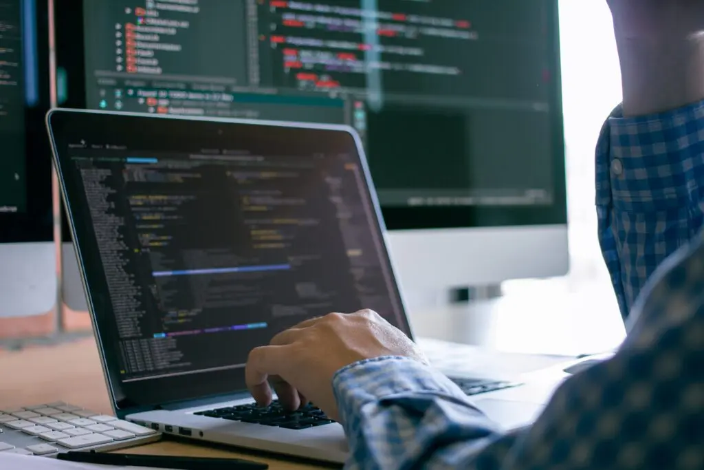 Person tippt auf einem Laptop mit Quellcode, im Hintergrund ein weiterer Monitor mit Code
