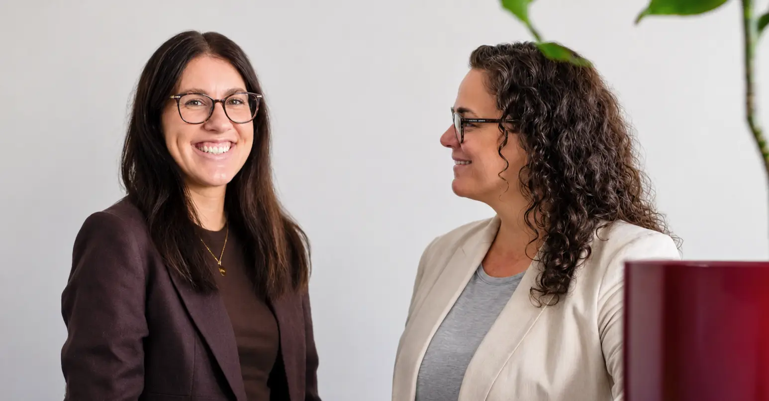 Zwei Frauen mit Brille, eine lächelt in die Kamera, die andere schaut seitlich, beide tragen Blazer und stehen vor einer weißen Wand mit einer grünen Pflanze im Vordergrund