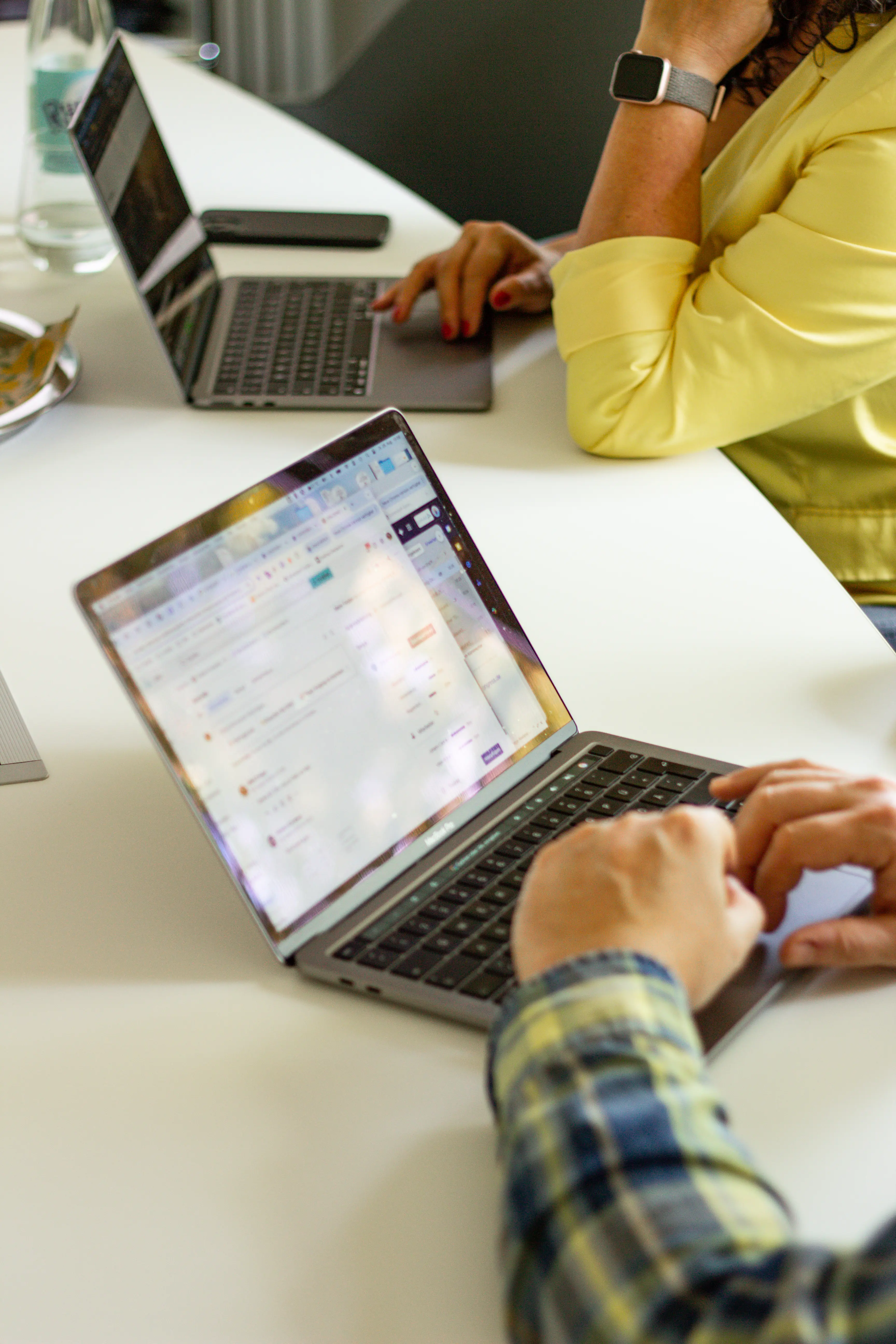 Zwei Personen arbeiten an Laptops an einem weißen Tisch, eine trägt ein gelbes Oberteil, die andere ein kariertes Hemd