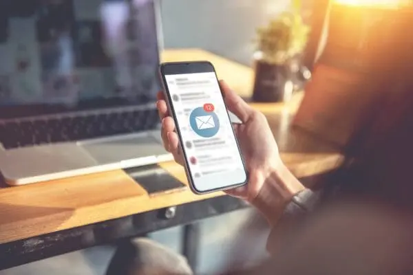 Hand hält Smartphone mit geöffneter E-Mail-App und Benachrichtigungssymbol vor Laptop auf Holztisch