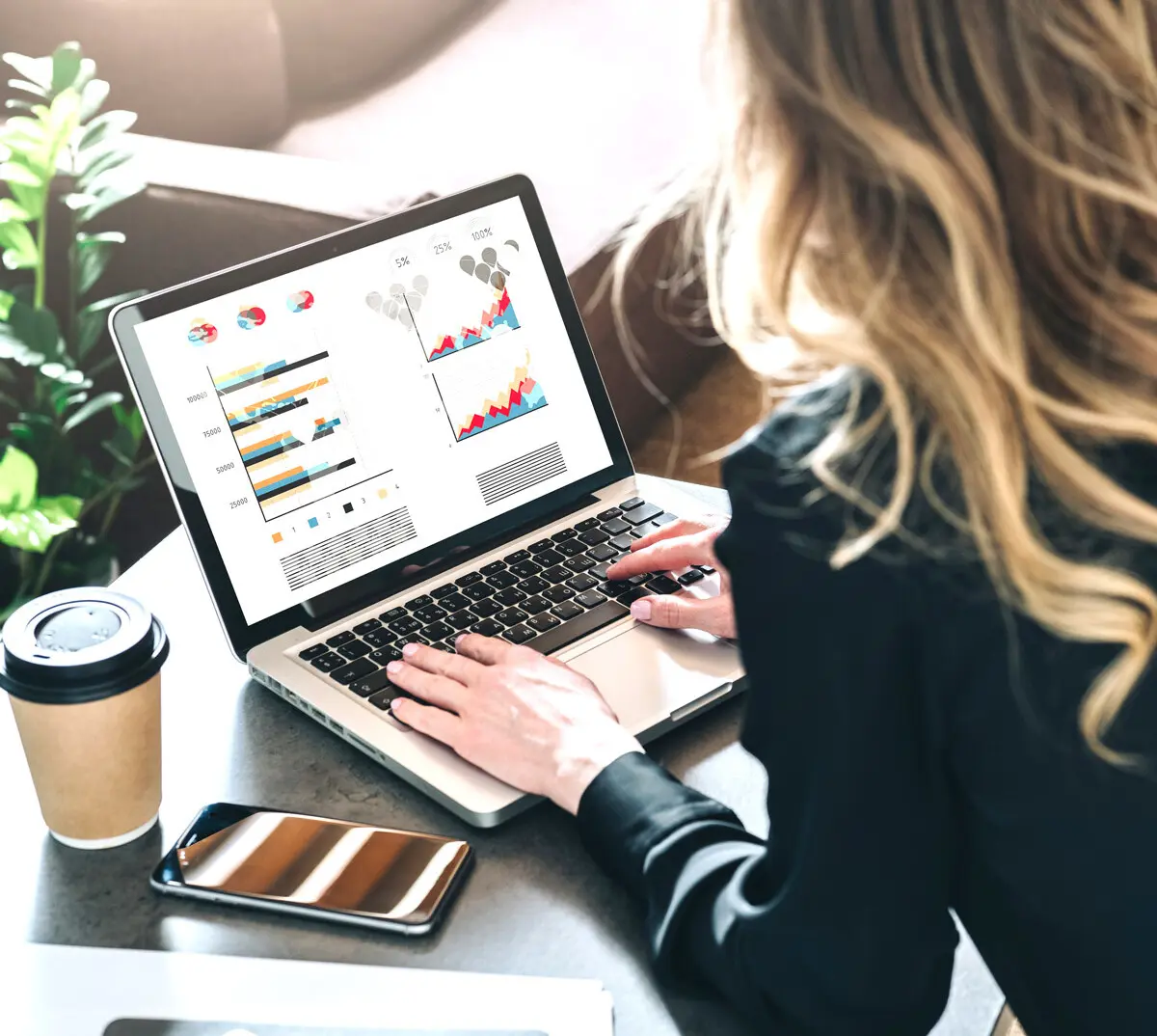 Frau tippt auf einem Laptop mit bunten Diagrammen und Statistiken auf dem Bildschirm, daneben ein Smartphone und ein Kaffeebecher