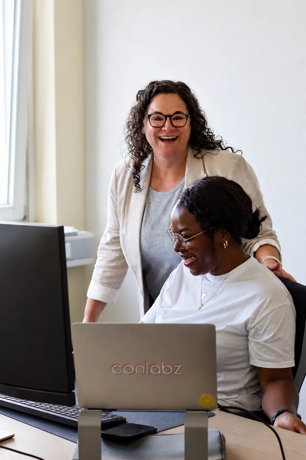 Zwei Frauen lachen gemeinsam vor einem Laptop mit conlabz-Logo – Teamarbeit Digitalagentur conlabz Koblenz
