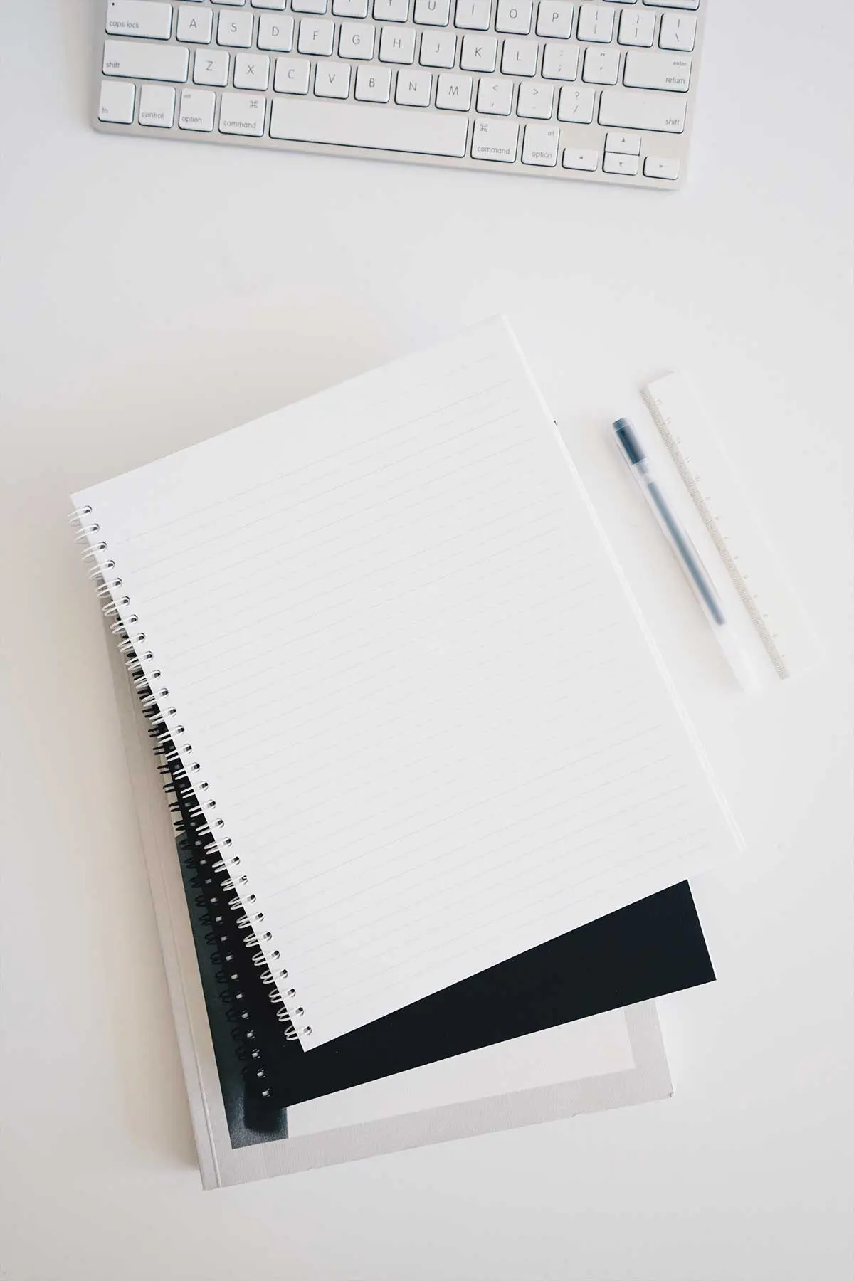 Ein aufgeschlagener, linierter Spiralblock liegt schräg auf einem Stapel Notizbücher auf einem weißen Schreibtisch. Rechts daneben befinden sich ein Stift und ein transparentes Lineal. Am oberen Bildrand ist der untere Teil einer silbernen Computertastatur angeschnitten.