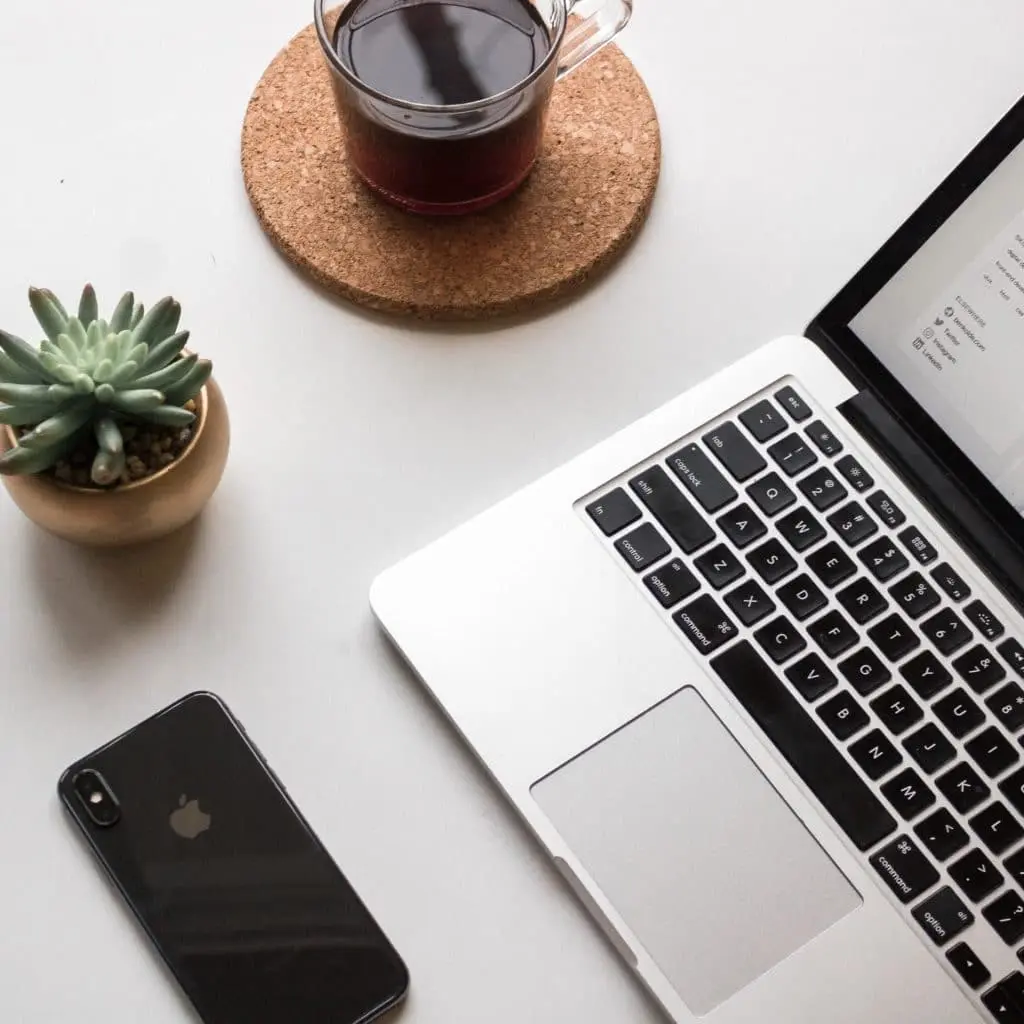 Draufsicht auf einen aufgeräumten, weißen Schreibtisch mit einem modernen Tech-Setup. Oben rechts steht ein geöffneter, silberner Laptop mit schwarzer Tastatur. Unten links liegt ein schwarzes iPhone mit sichtbarem Apple-Logo. Oben links steht eine Sukkulente in einem runden Goldtopf. Oben mittig befindet sich eine Glastasse Kaffee auf einem runden Korkuntersetzer.