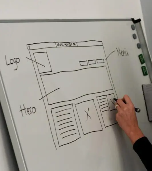 Hand zeichnet mit einem Stift ein Website-Wireframe auf ein Whiteboard, beschriftet mit Logo, Menü und Hero