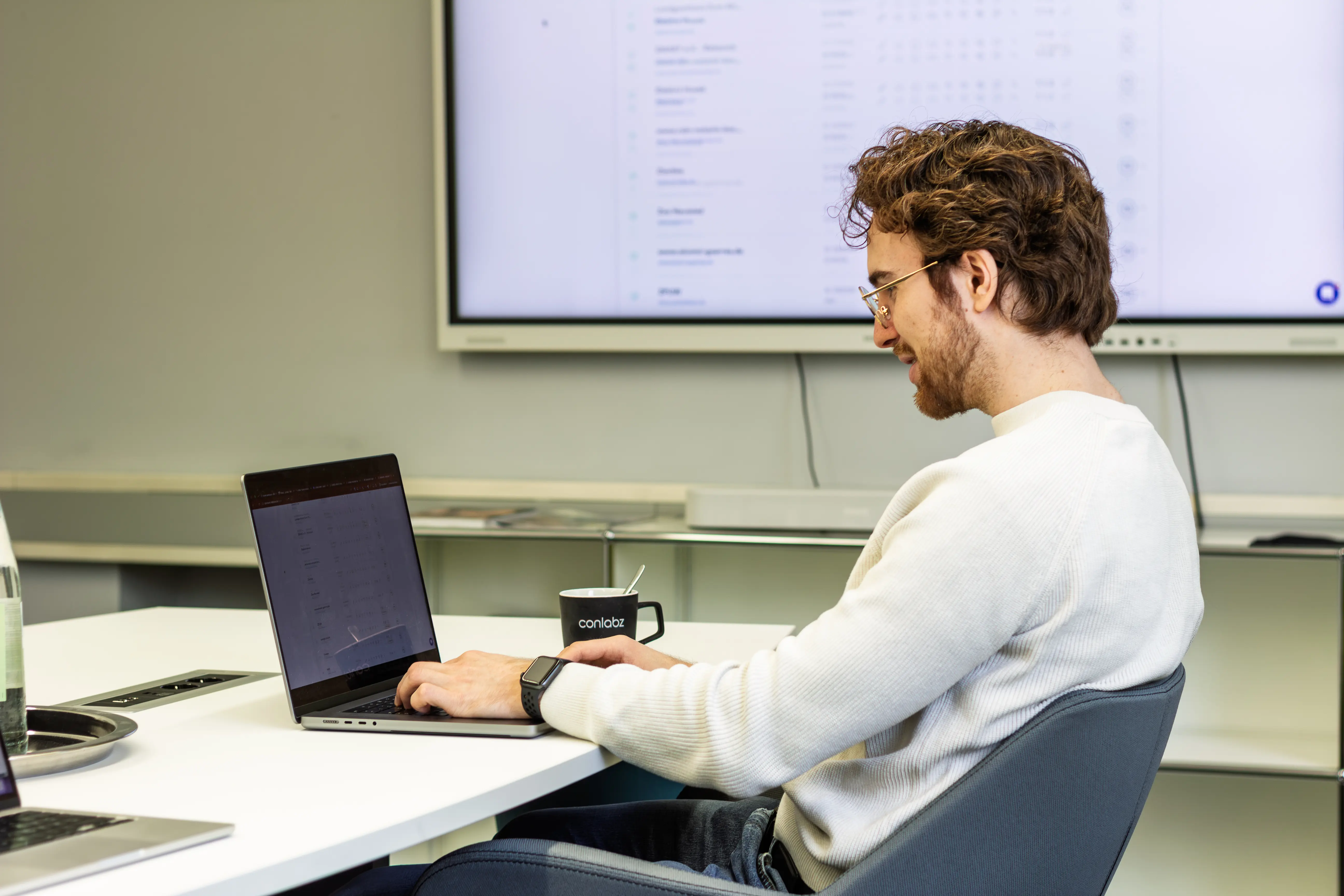 Entwickler arbeitet konzentriert am Laptop, im Hintergrund ein Monitor mit einer Systemübersicht